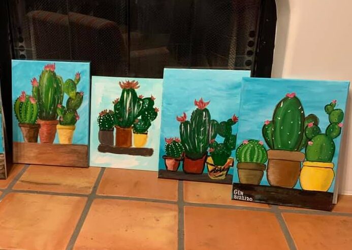 Five paintings of cacti in pots are displayed in front of a fireplace with glass doors. The paintings are colorful, featuring various types and sizes of cacti with some blooming pink flowers, set against blue sky backgrounds. They rest on a tiled floor.