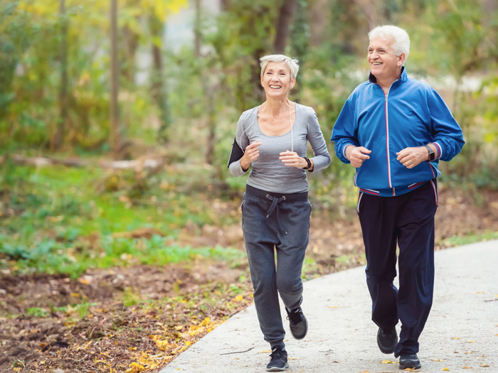 A senior couple jogs together on a paved path through a lush, green park. The woman, wearing a gray workout outfit, smiles while jogging beside the man, who is dressed in a blue jacket and black pants. Both appear happy and energetic amidst the natural surroundings.