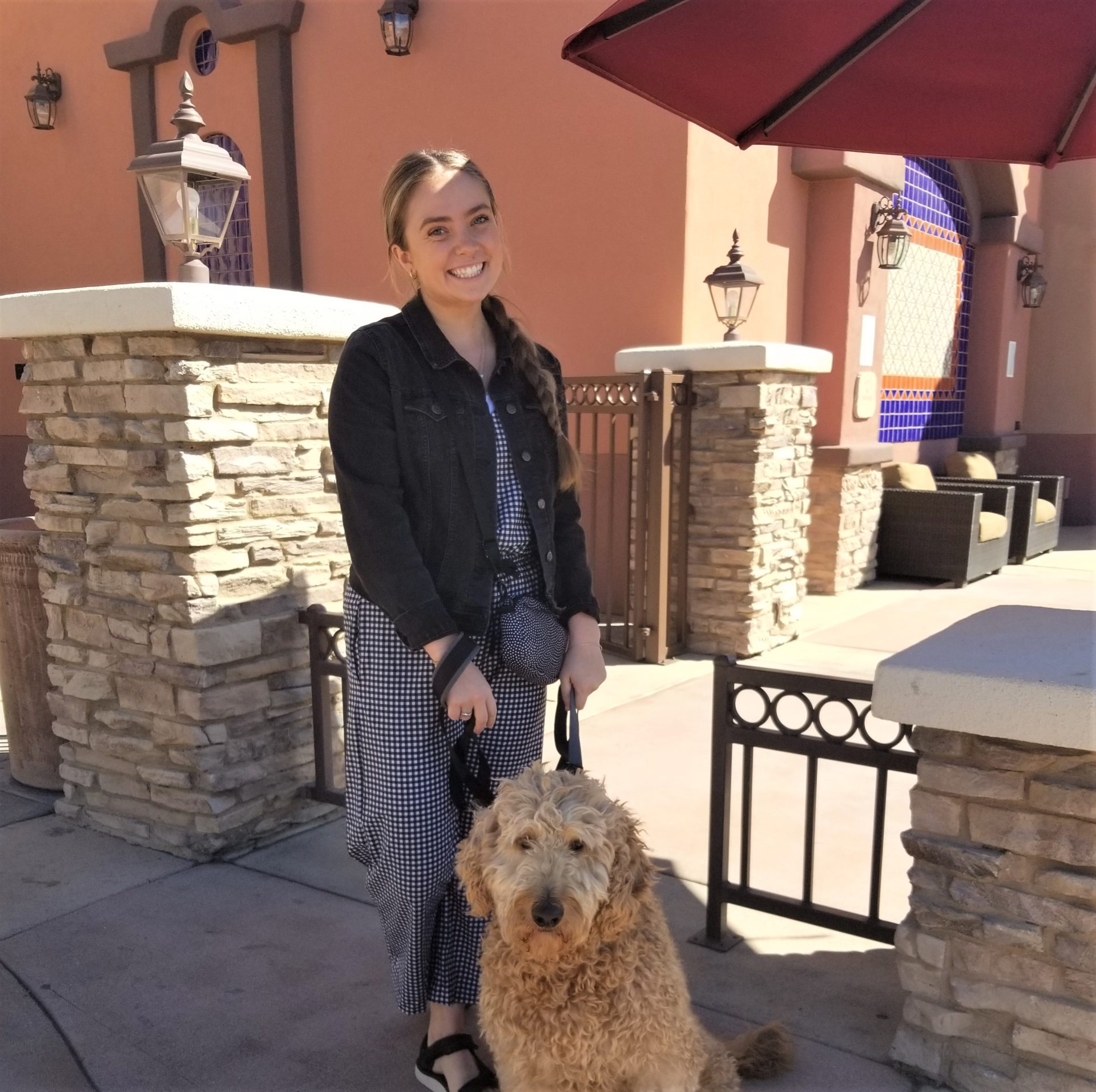 A woman smiles while standing under a red umbrella and holding a fluffy, curly-haired dog by its leash. She is wearing a black jacket over a checkered outfit. The background shows a building with stone columns and outdoor seating.