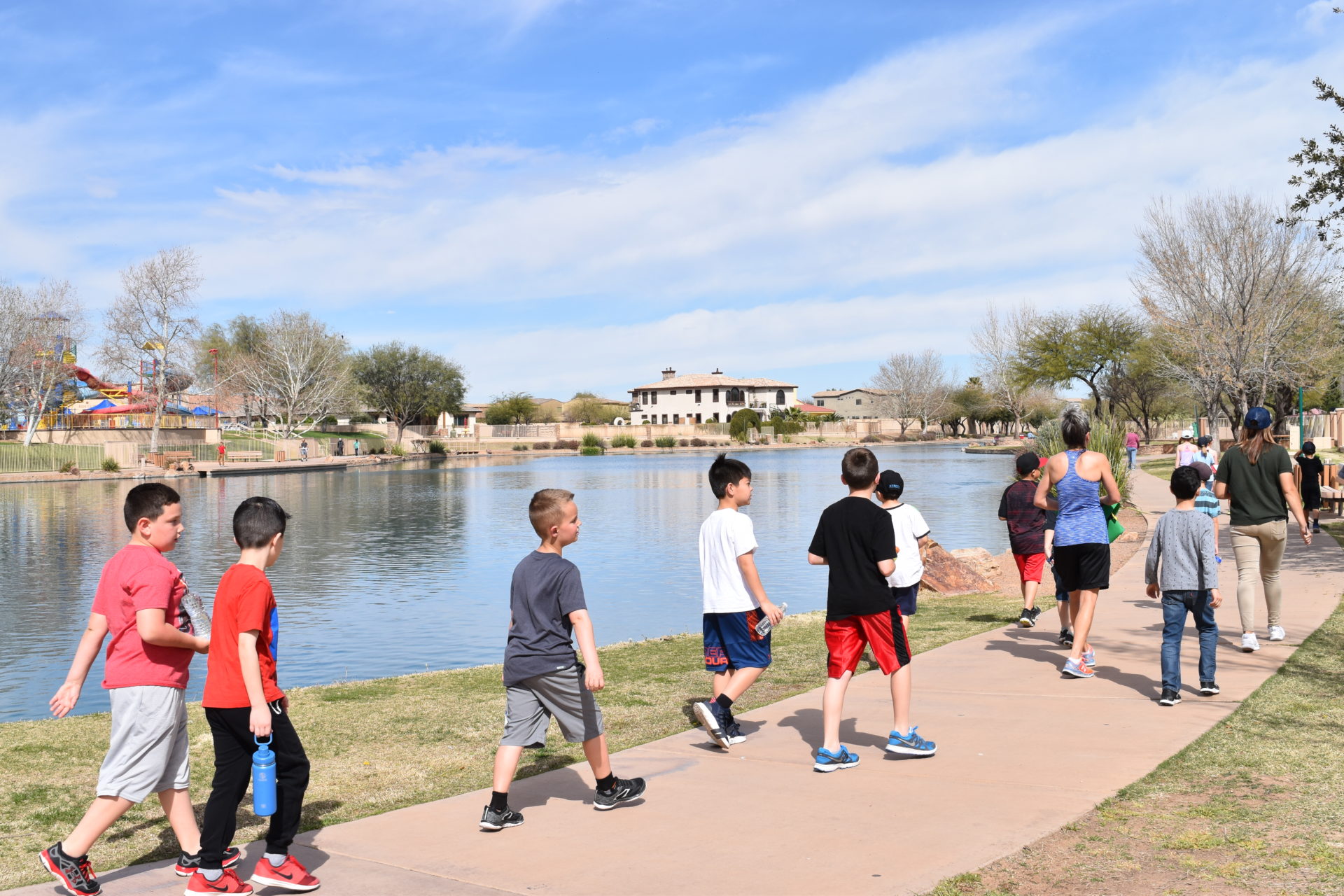 Children and adults walk along a paved path next to a large pond in a park on a sunny day. Trees and a building are visible in the background, with a playground off to the side. The sky is clear with a few clouds.