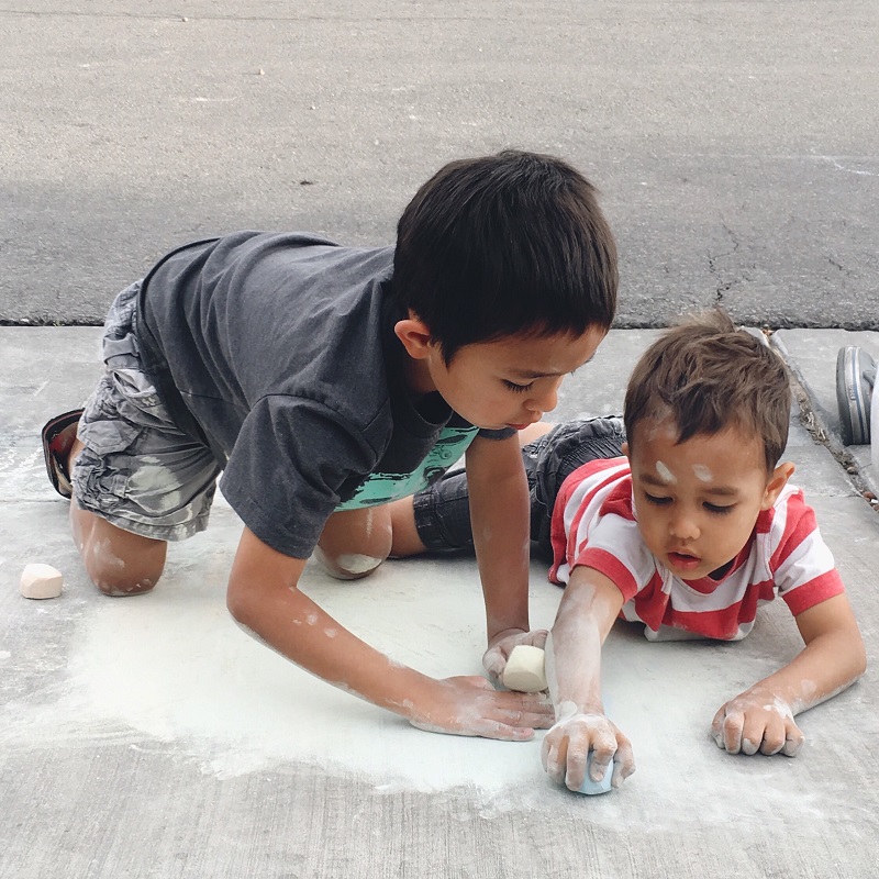 Two young children are kneeling and sitting on a concrete sidewalk, drawing with large pieces of chalk. The older child, wearing a grey shirt and patterned shorts, focuses on his artwork, while the younger child, in a red and white striped shirt, draws nearby.