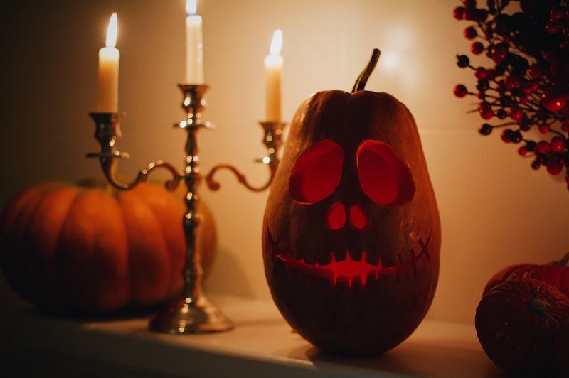 A carved pumpkin with a smiling face lit from within is situated next to a silver candelabra holding three lit candles. Another pumpkin and some autumnal decorations are placed around it, creating a cozy, warm ambiance.