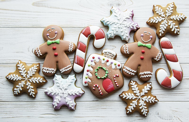 An assortment of festive gingerbread cookies arranged on a white wooden surface. The cookies include gingerbread men, candy canes, snowflakes, and a house decorated with colorful icing. Some of the decorations are in red, green, white, and purple.