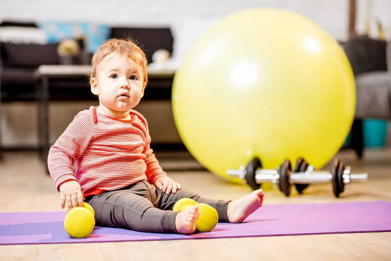 A baby wearing a red striped shirt and gray leggings sits on a purple exercise mat indoors, holding a small yellow ball in each hand. In the background, there's a large yellow exercise ball and a set of dumbbells.