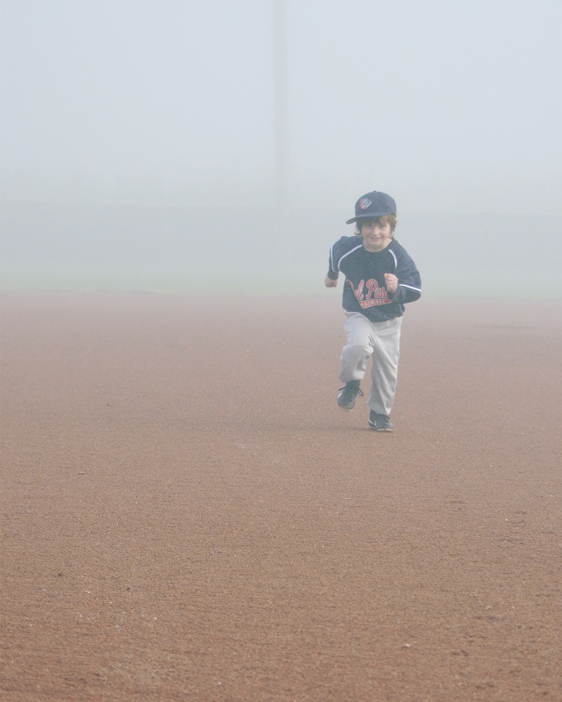 A young child in a baseball uniform runs enthusiastically across a foggy baseball field. The child is wearing a hat, a dark long-sleeve jersey, white pants, and cleats, smiling joyfully with one leg lifted in mid-stride. The dense fog obscures the background.