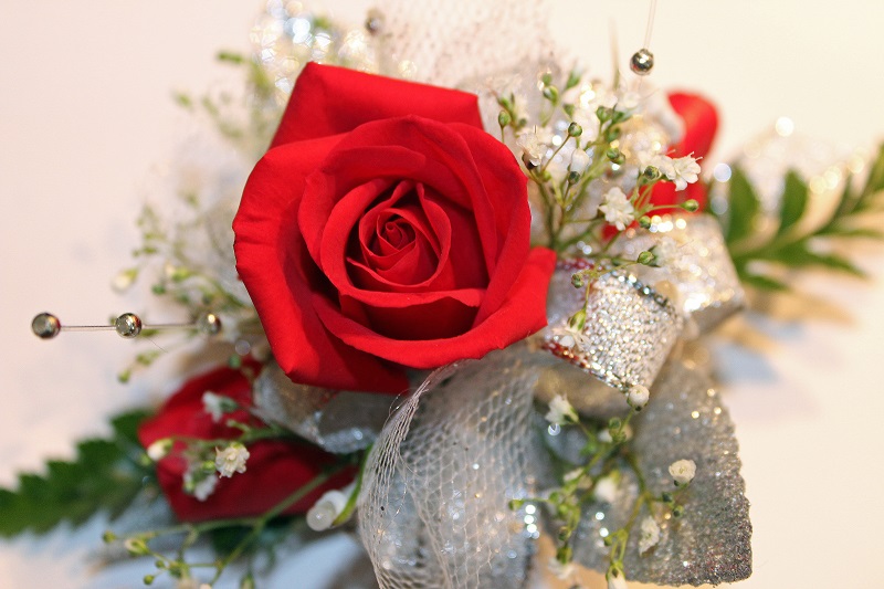 A close-up of a vibrant red rose surrounded by sprigs of baby's breath and greenery. The arrangement is embellished with sparkling silver mesh and decorative pins, creating an elegant and festive appearance against a light background.