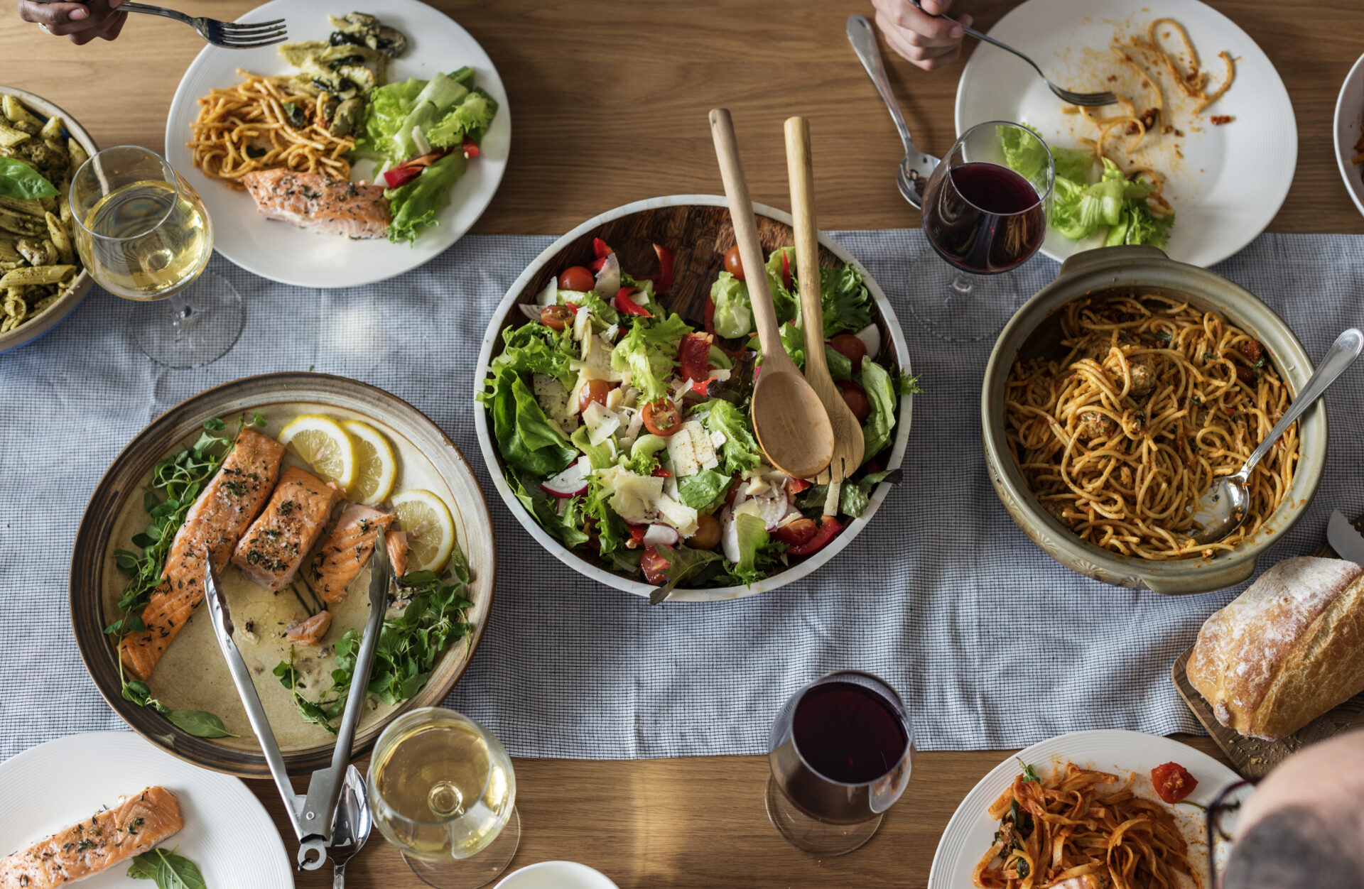 A wooden table set for a meal with several dishes. In the center, a large bowl of fresh salad with wooden serving utensils. Surrounding it are plates of pasta, salmon with lemon, and a half-eaten dish. There are also glasses of red and white wine.