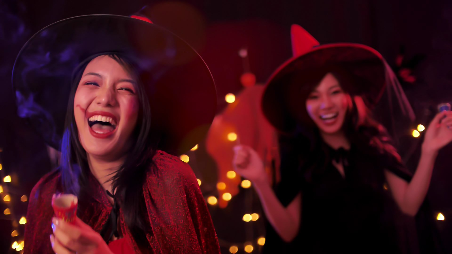 Two women, dressed in Halloween costumes with witch hats, are laughing and having a good time. The woman in the foreground has a painted face and is holding a beverage. The background is adorned with warm, festive lights.