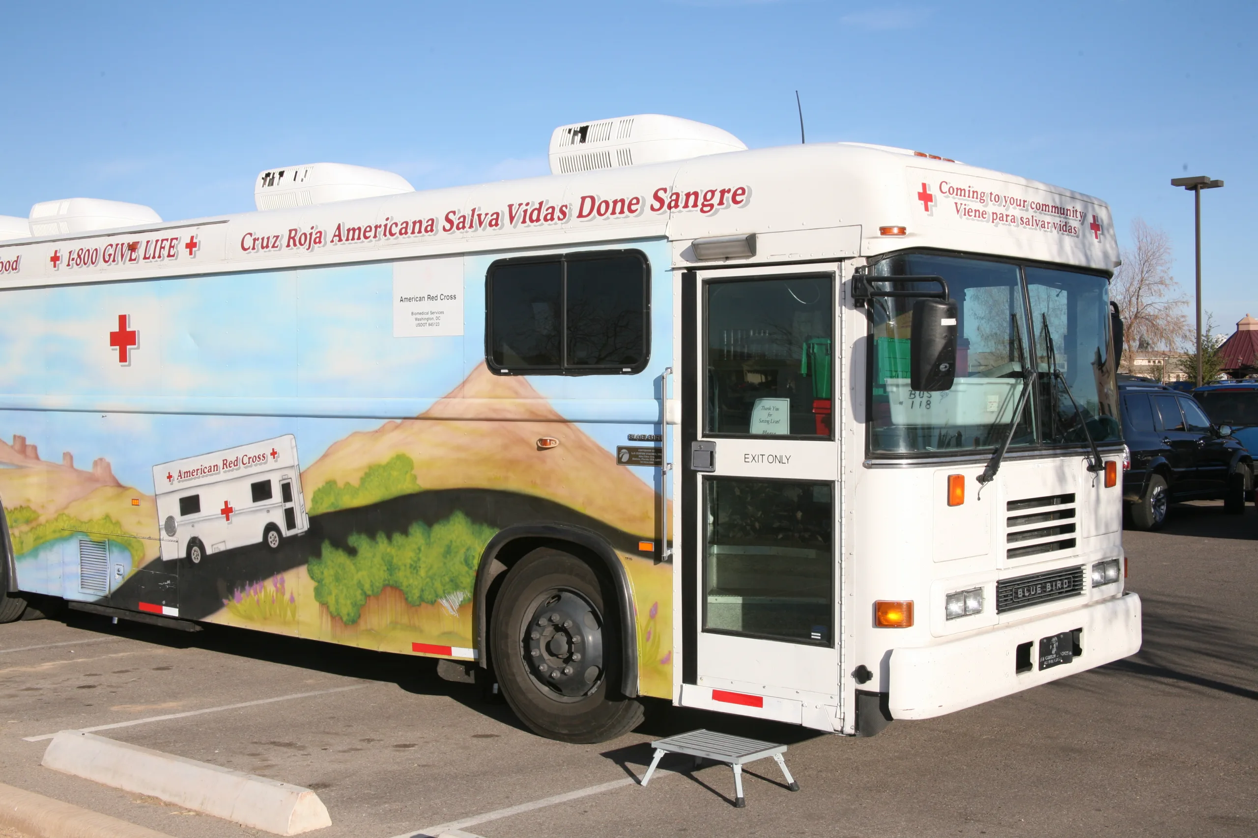 A large white mobile blood donation bus with Red Cross logos and Spanish text that translates to "American Red Cross Saves Lives. Donate Blood." The bus is parked in a parking lot on a sunny day, with a small set of steps leading up to its entrance.