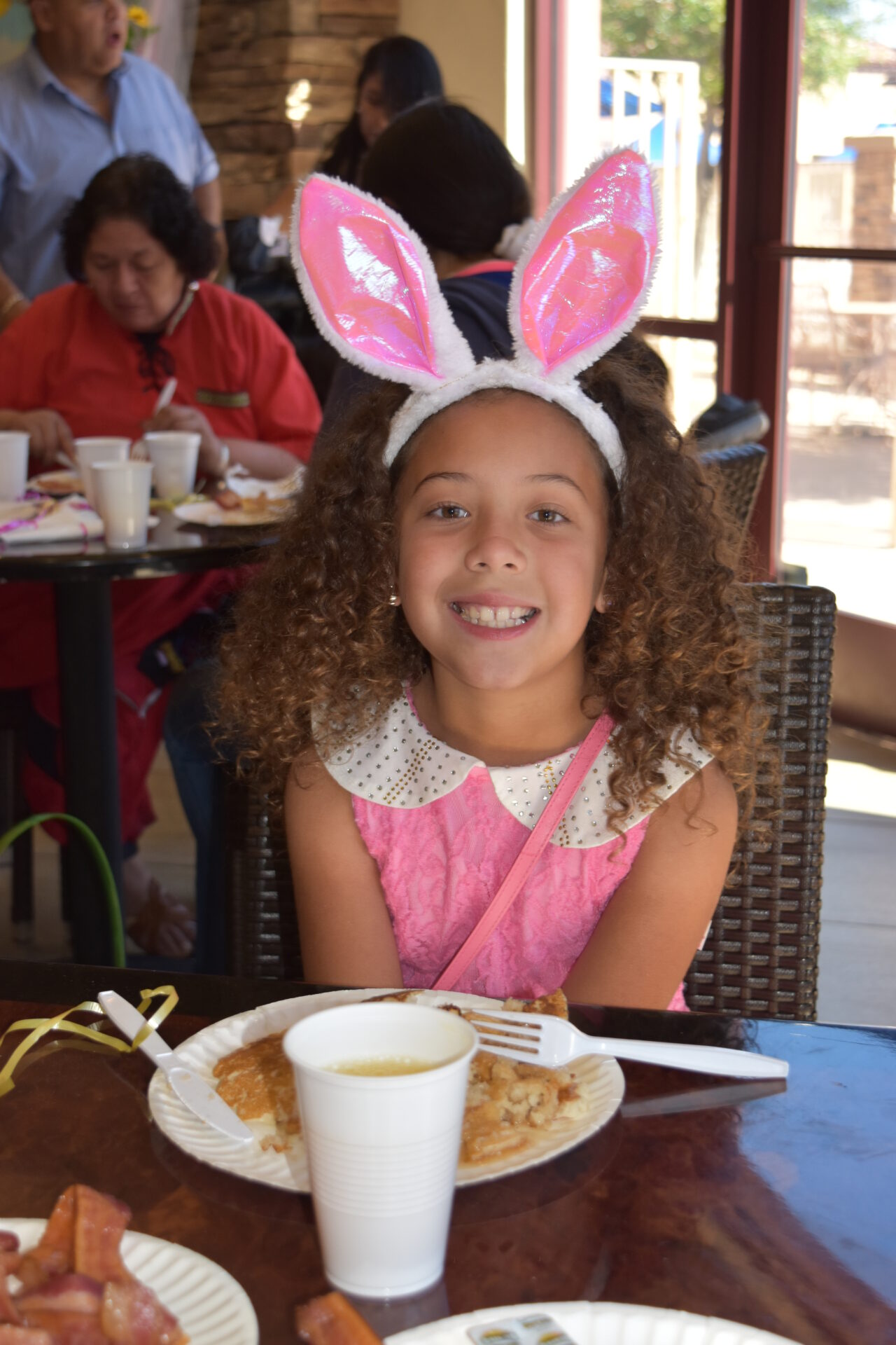 A young girl with curly hair, wearing a pink dress and bunny ears headband, smiles while seated at a table with pancakes and bacon on a plate. In the background, there are other people sitting and eating at the table.