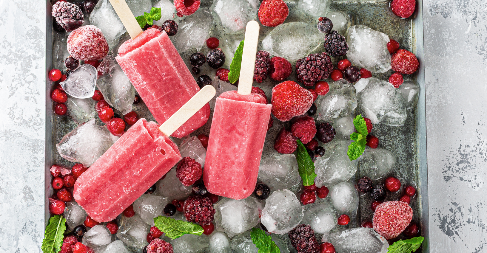 Three pink popsicles on wooden sticks are placed on a bed of ice cubes and a variety of frozen berries, including strawberries, blackberries, raspberries, and red currants. Mint leaves are scattered around for garnish, adding a touch of green to the icy scene.