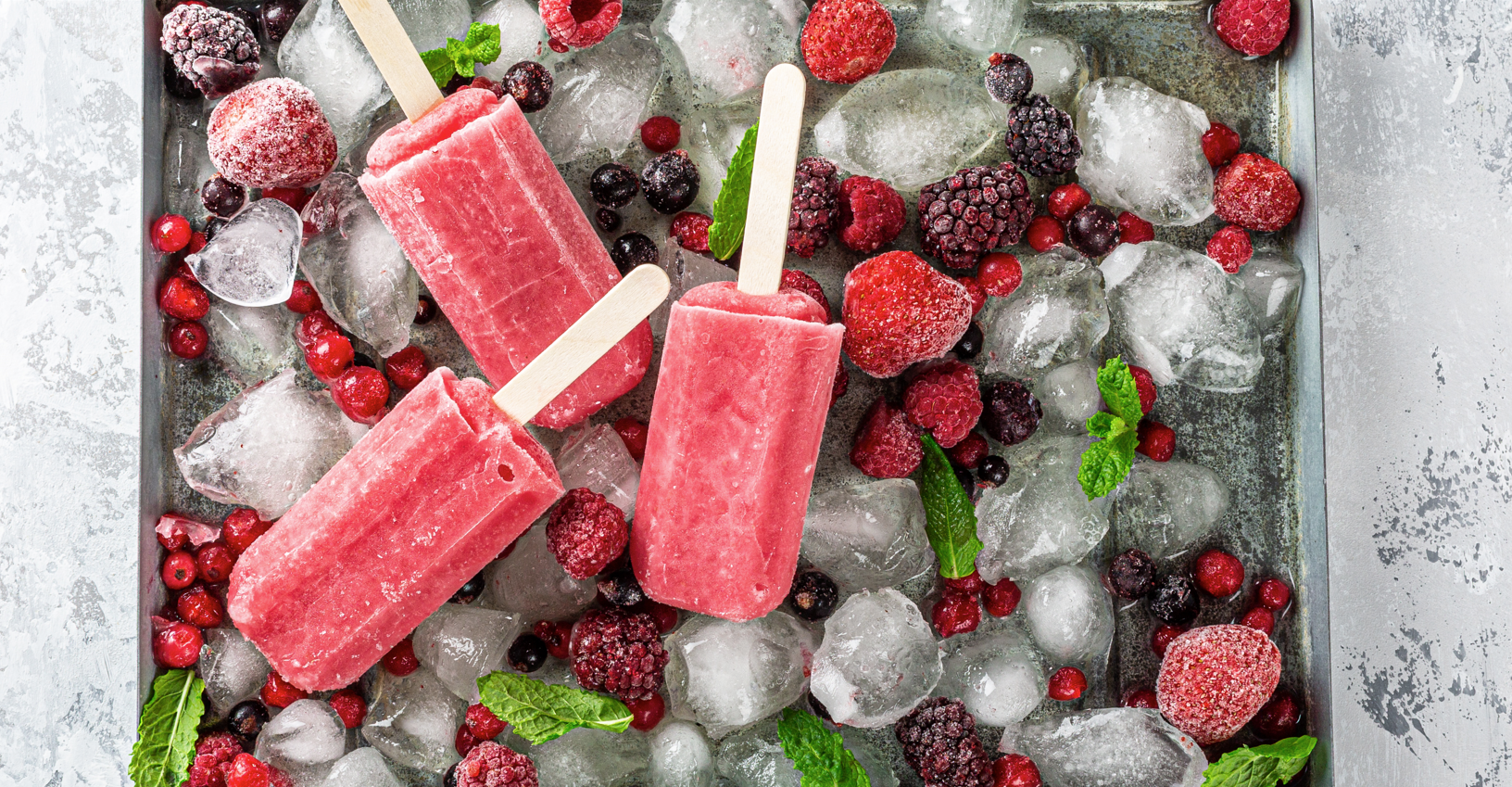 Three pink frozen fruit popsicles on wooden sticks are laid on a bed of ice cubes, surrounded by various frozen berries including raspberries, blackberries, and red currants. Green mint leaves are scattered among the berries and ice for garnish.