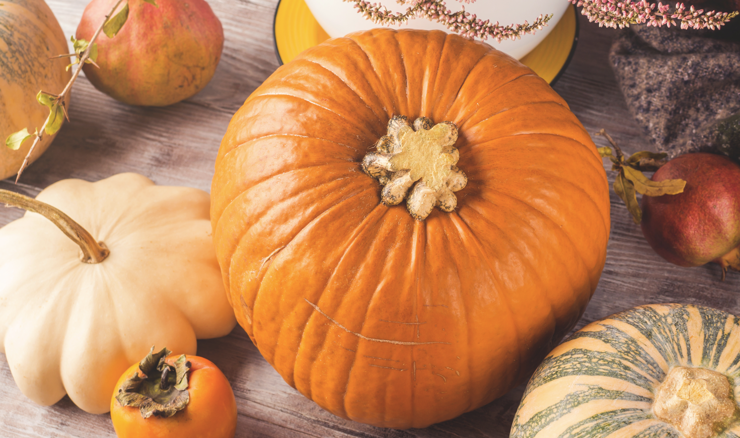 A wooden surface is adorned with an array of autumn produce. The centerpiece is a large, orange pumpkin with an off-white stem. Surrounding it are a white pumpkin, a green-striped squash, a persimmon, a red pomegranate, and some decorative foliage.