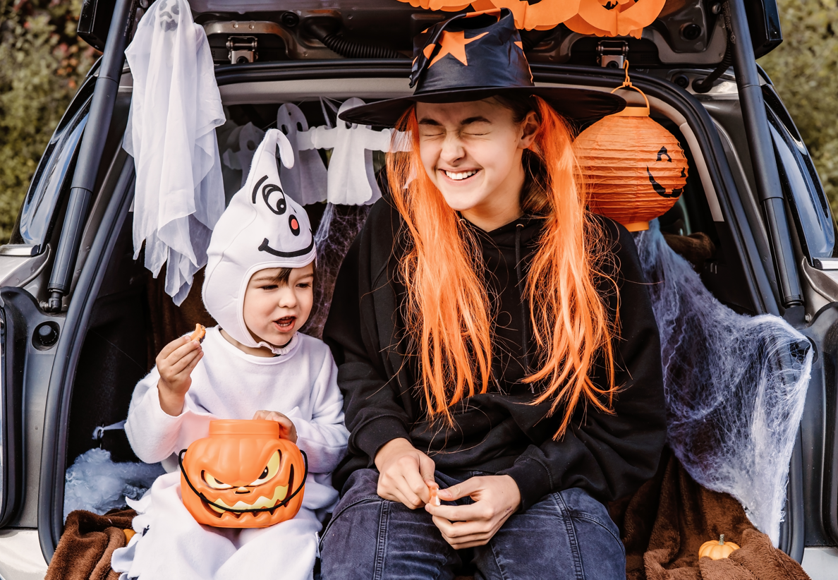 A cheerful child and an adult, both in Halloween costumes, sit in the back of a car decorated with Halloween decorations. The child is dressed as a ghost and holds a pumpkin-shaped bucket, while the adult wears a witch hat and has long orange hair.