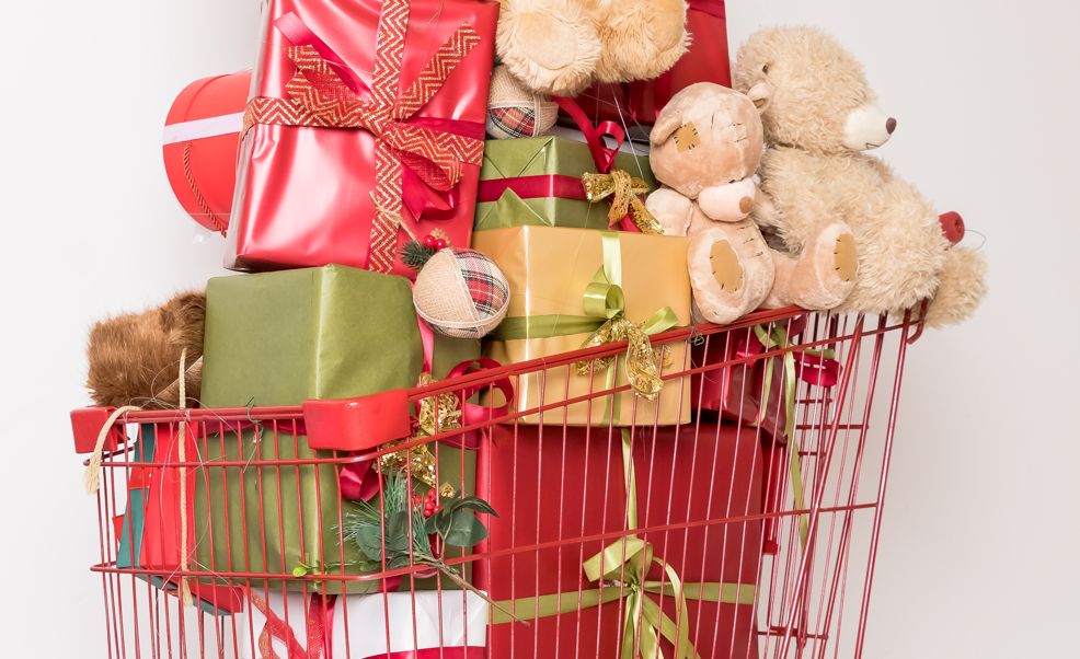 A red shopping cart overflowing with Christmas presents and stuffed animals. The gifts are wrapped in various festive papers and adorned with ribbons. The stuffed animals include multiple teddy bears, all arranged amidst the colorful packages.