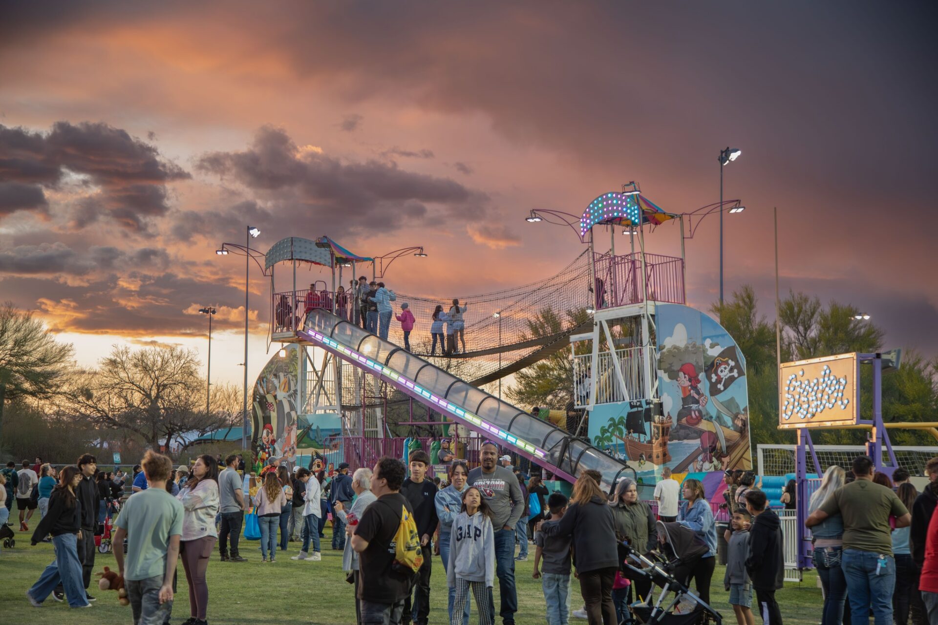 A lively crowd enjoys a fair at sunset, with people walking, talking, and kids playing near a large, colorful slide structure decorated with cartoon art under a dramatic, cloudy sky.