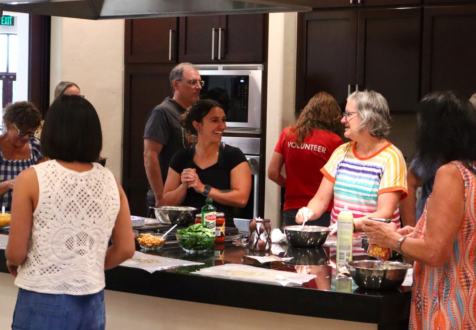 A group of people gather around a kitchen island, preparing food and smiling. Various bowls and ingredients are on the counter. The setting appears lively and communal, with participants engaged in conversation.