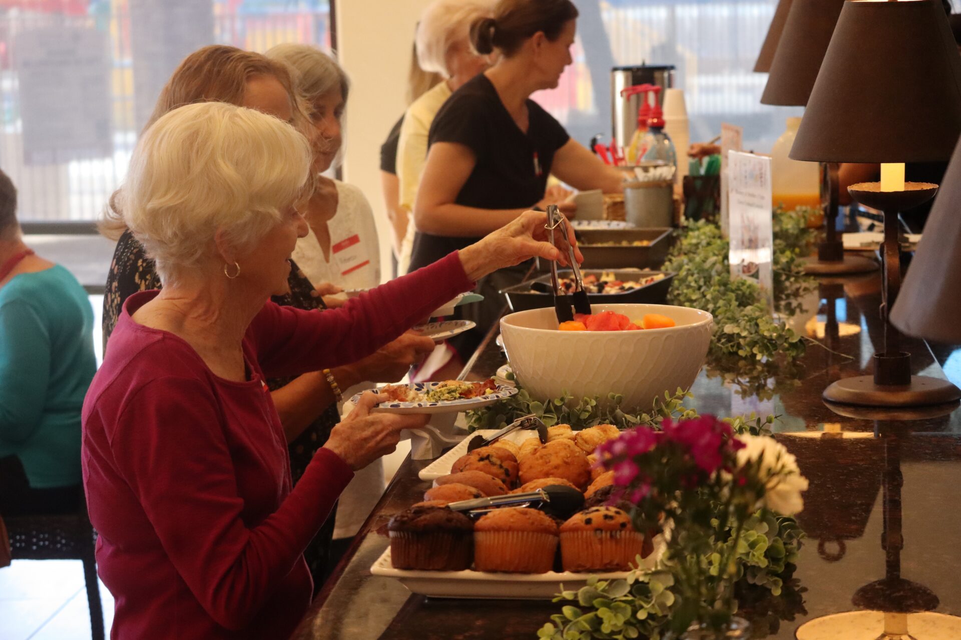Older women serve themselves food from a buffet table, selecting salad and muffins. The setting appears social and relaxed, with flowers and greenery decorating the counter.