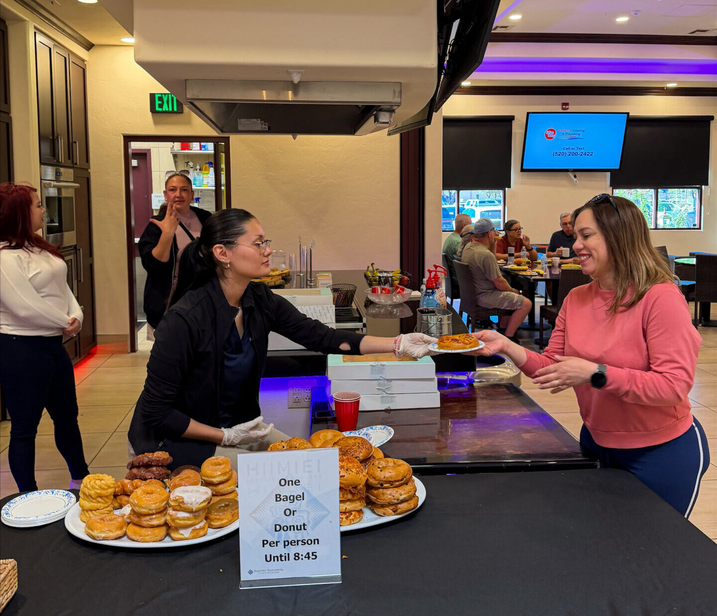 A woman hands a bagel to another woman at a table with stacked bagels and pastries. A sign reads, "One Bagel Or Donut Per Person Until 8:45." People are standing and chatting in a bright indoor setting.