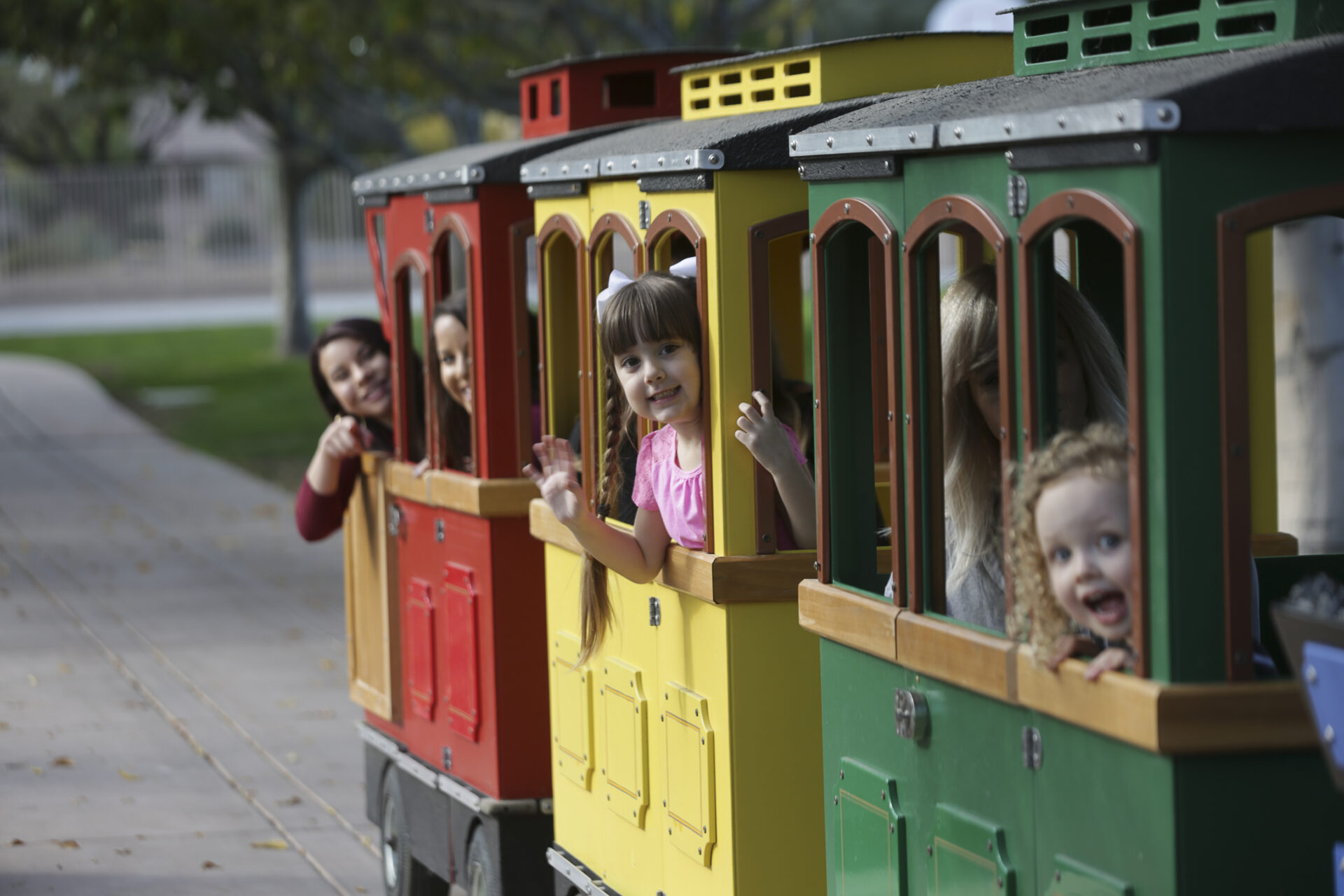 Children and adults smile and wave from the windows of colorful red, yellow, and green train cars on an outdoor track, with trees and a sidewalk in the background.