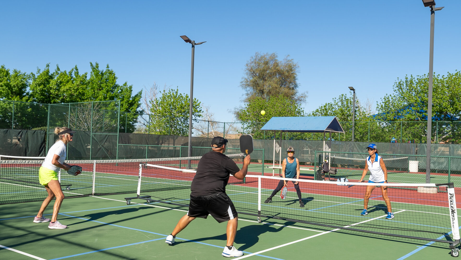 DSC01346 Four adults play pickleball outdoors on a sunny day, showcasing why this is the best place to live in Arizona. Two women and two men are ready with paddles by the net, as one prepares to hit the ball. Lush trees and fencing frame the scene.