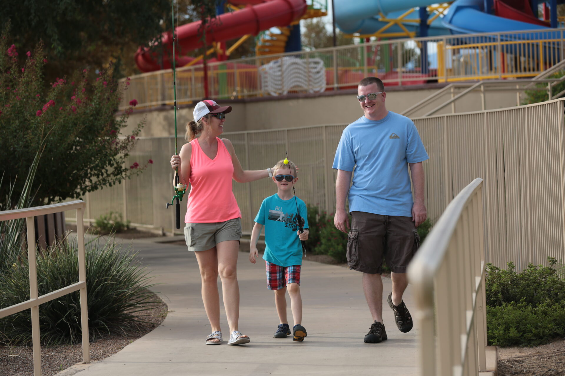 DSPH3856 A woman, a man, and a boy walk together outside near a fence. The woman holds a fishing rod and playfully touches the boy’s head, while the man smiles. Water slides are visible in the background.