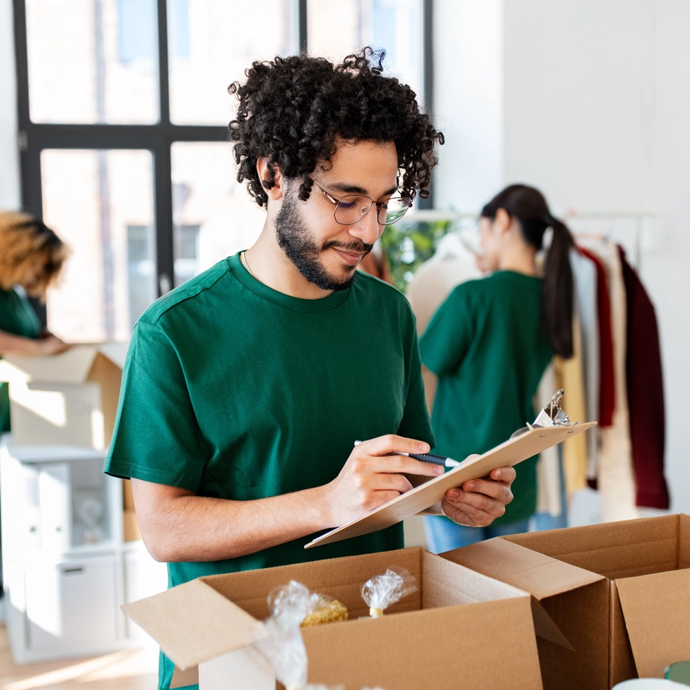 person-packing-donations-and-writing-on-a-clipboard A man in a green shirt writes on a clipboard while standing next to open cardboard boxes. Other people pack items into boxes in the background in a bright, spacious room.