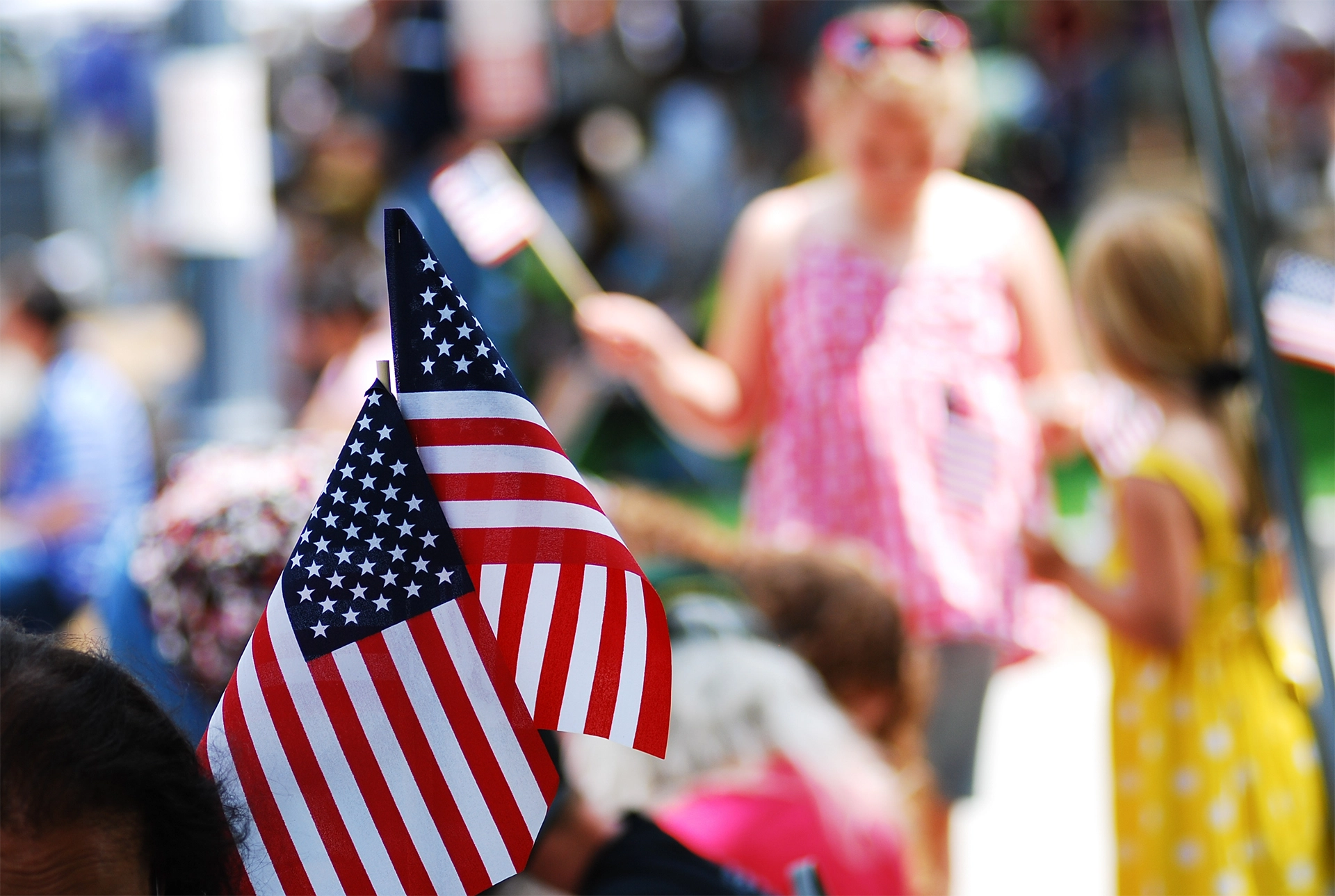 Close-up of small American flags with blurred people in summer clothing, including children holding flags, in the background at an outdoor patriotic event.