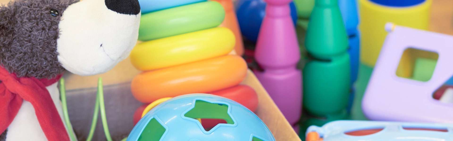 teddy-bear-and-toys-in-a-pile A close-up of colorful children’s toys, including a plush bear with a red scarf, stacking rings, geometric shapes, and a blue shape sorter ball, all arranged together on a table.