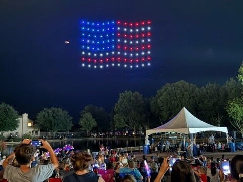 A crowd watches as a formation of drones lights up the night sky in the shape and colors of the American flag above a park, with trees and a white tent visible in the foreground.
