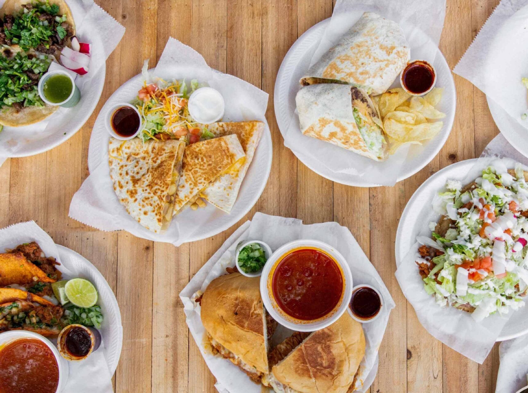 An overhead view of various Mexican dishes on paper plates, including quesadillas, sandwiches, tacos, salad, chips, and sauces, displayed on a wooden table. Uber Eats logo appears in the bottom right corner.
