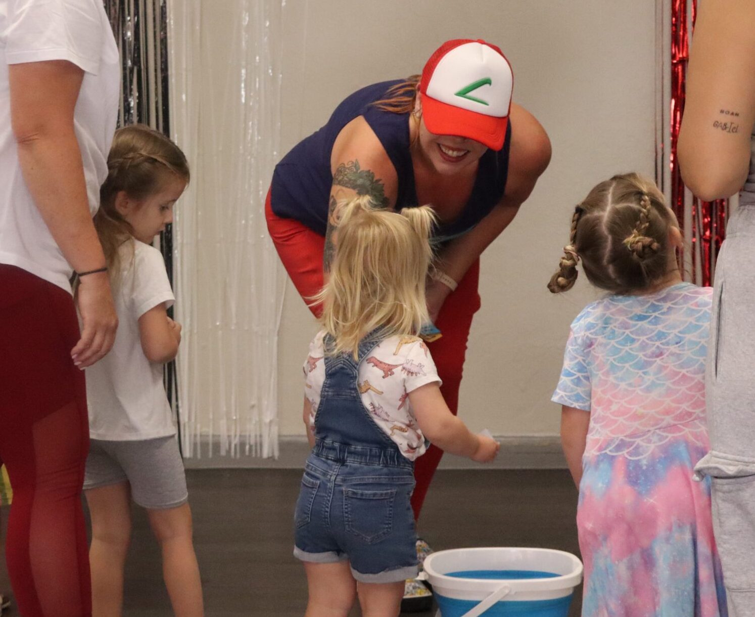A woman in a red cap bends down, smiling and talking to young children gathered around a bucket indoors. The children are dressed casually, and the atmosphere is cheerful and playful.