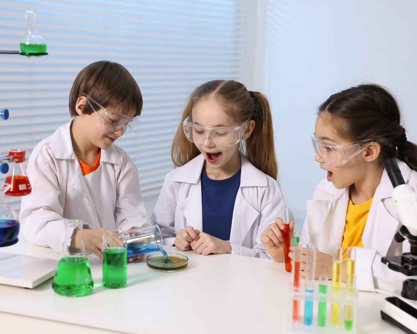 Three children in lab coats and safety goggles conduct a science experiment with colorful liquids and a petri dish, appearing excited and engaged in a bright laboratory setting.