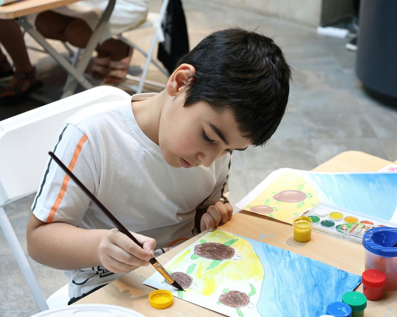 A young boy with dark hair sits at a table painting a picture with yellow, green, and brown colors. Art supplies, including paint pots and brushes, are on the table beside him. He is focused on his artwork.