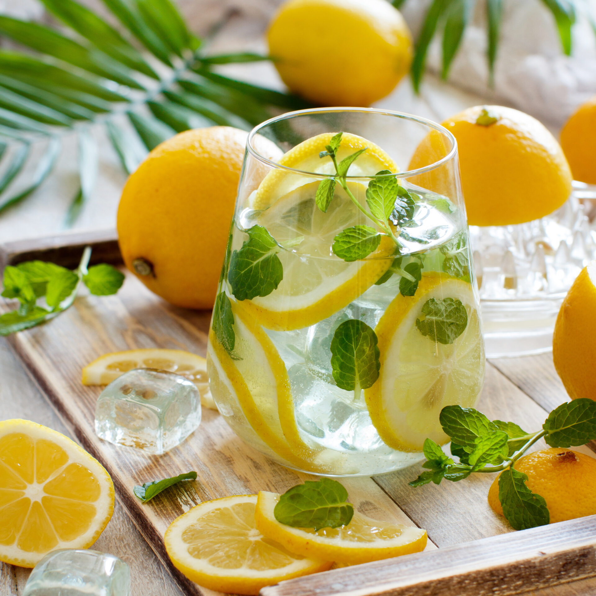 A refreshing drink with lemon slices, mint leaves, and ice cubes in a glass on a wooden tray, surrounded by whole lemons, lemon slices, and mint sprigs.