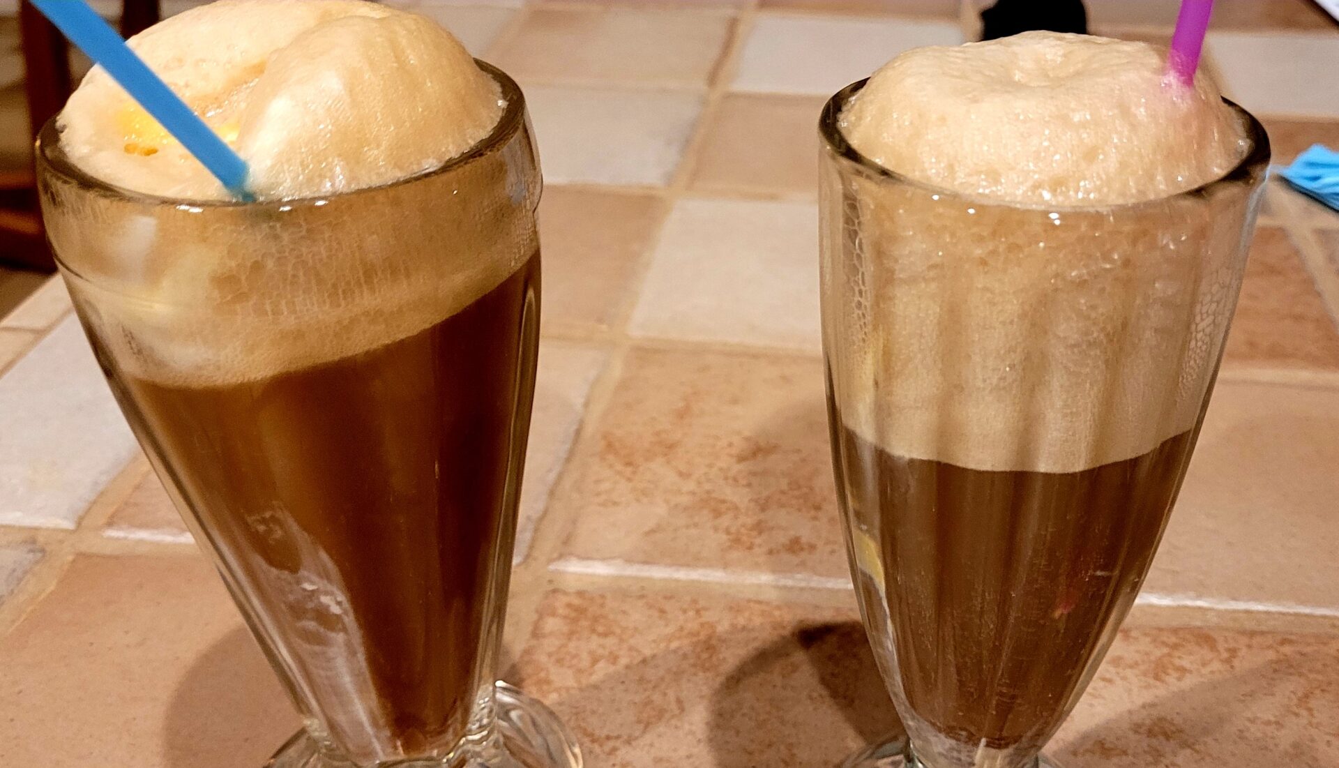 Two tall glasses filled with root beer floats, topped with foamy vanilla ice cream, each with a colorful straw, sit on a tiled kitchen counter.