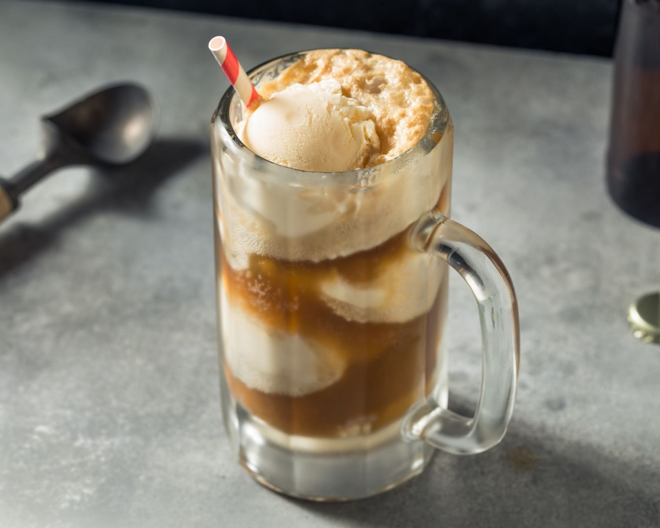A glass mug filled with root beer and scoops of vanilla ice cream, creating a root beer float. A red and white striped straw is in the mug, and there is a metal spoon in the background on a gray surface.