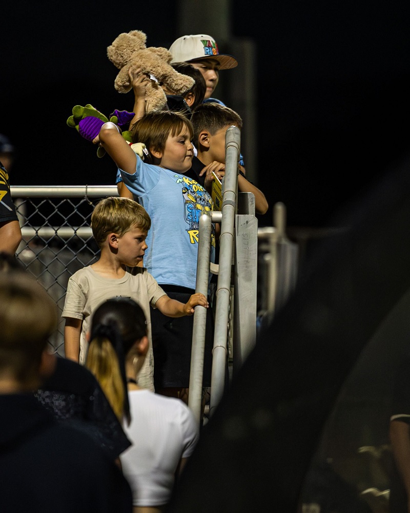 A group of children stand in stadium bleachers at night, with one boy in a blue shirt excitedly waving a teddy bear above his head. Other kids and adults watch the event, some leaning on the railings.