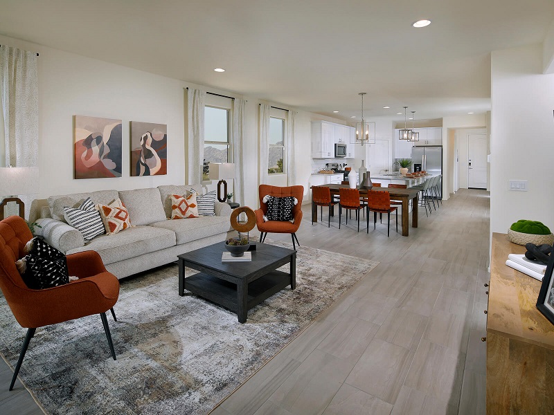 Modern open-concept living and dining area with light wood floors, a beige sofa, orange chairs, abstract wall art, and a large dining table with orange chairs. The kitchen is visible in the background.