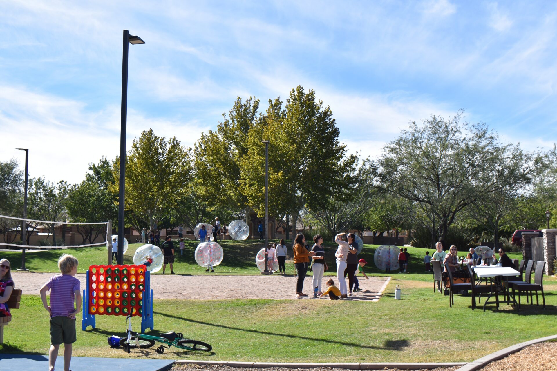 People enjoy outdoor games in a park, including giant Connect Four and bubble soccer. Trees, tables, and a volleyball net are in the background under a sunny sky. Children and adults are participating in activities.