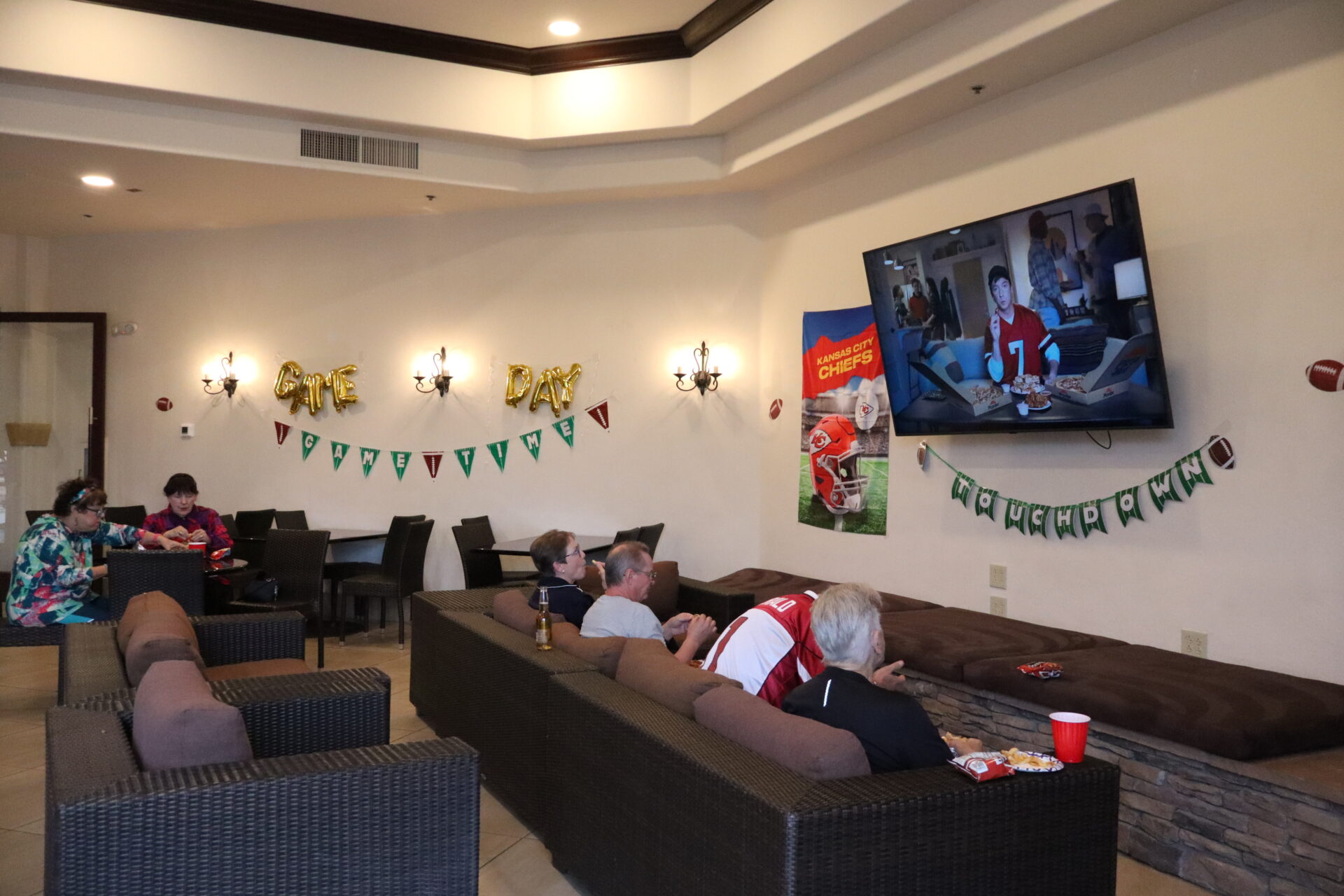 A group of people sit on couches in a lounge decorated with football-themed banners, watching a football game on a large wall-mounted TV. Food and drinks are on the tables. "Game Day" is spelled out on the wall.