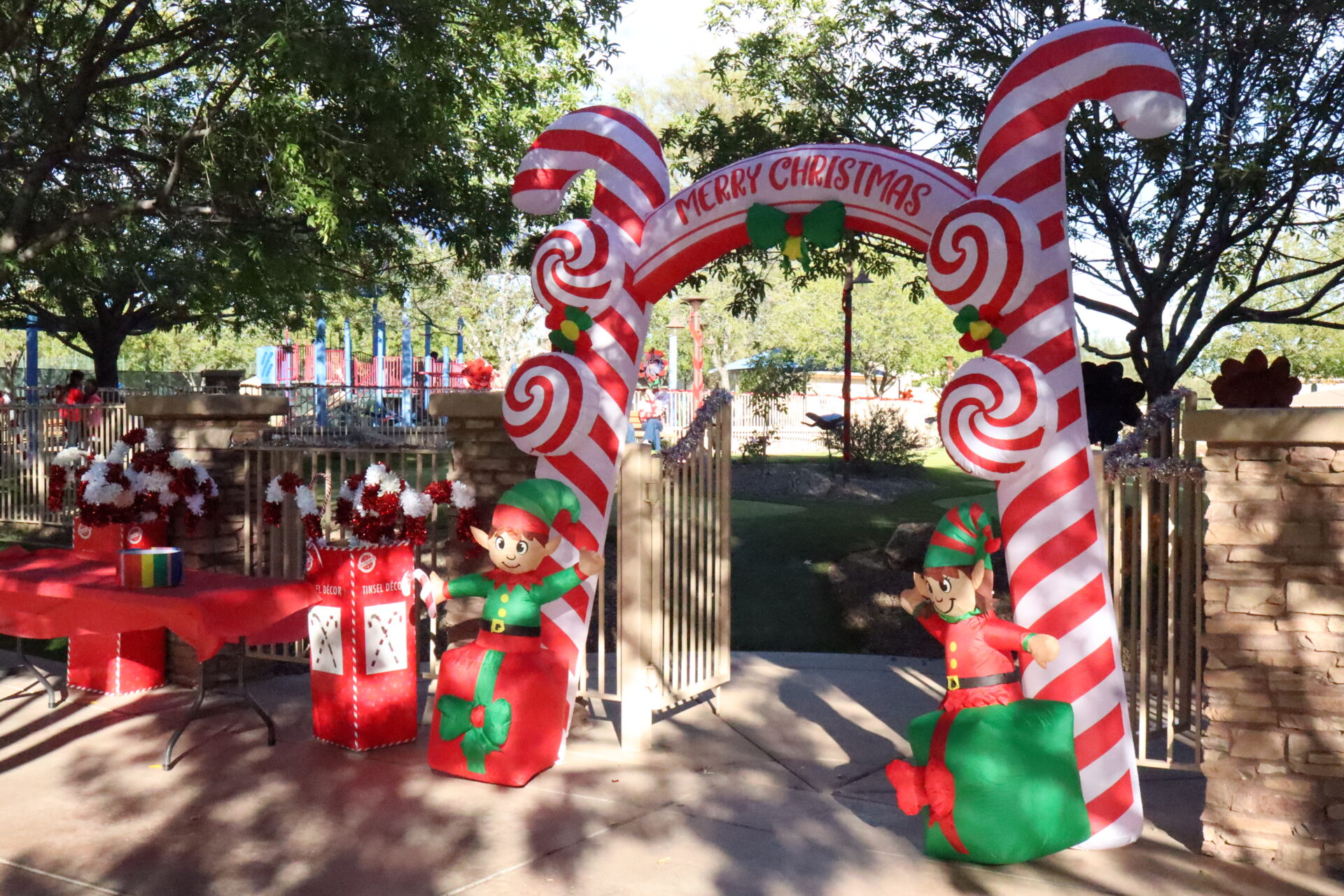 An inflatable candy cane archway labeled “Merry Christmas” featuring two toy elves sitting on presents, positioned at an entrance gate in a park.