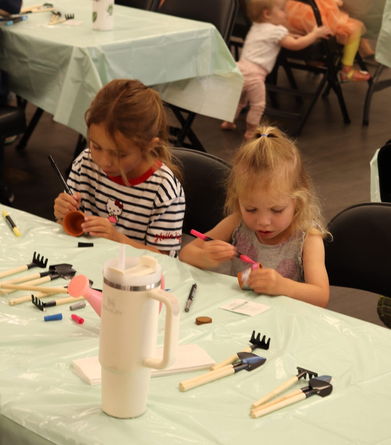 Two young children sit at a table covered with a light green tablecloth, painting small objects. Art supplies and water bottles are on the table. In the background, other children and adults are seated at similar tables.