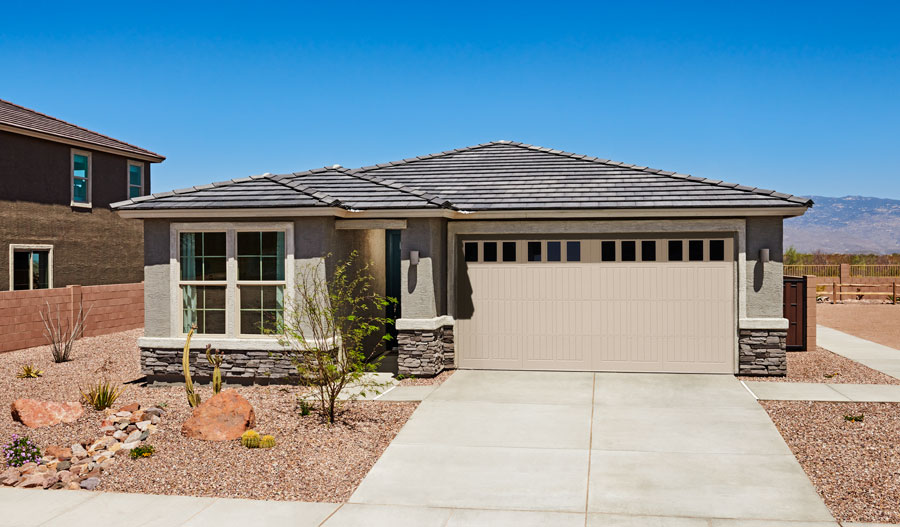 Single-story modern house with gray exterior, stone accents, and a two-car garage. The front yard features desert landscaping with rocks and drought-tolerant plants under a clear blue sky.