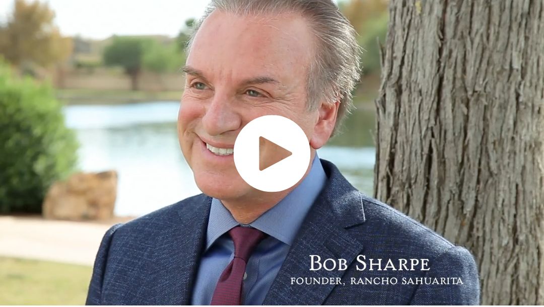 A man in a blue suit and red tie smiles while sitting outdoors near a tree and a lake. The text on the image reads, "Bob Sharpe, Founder, Rancho Sahuarita." A play button is centered on the image.