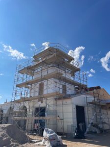 A multi-story building under construction, surrounded by scaffolding, with clear blue sky and a few clouds in the background. Construction materials and sand piles are visible in the foreground.