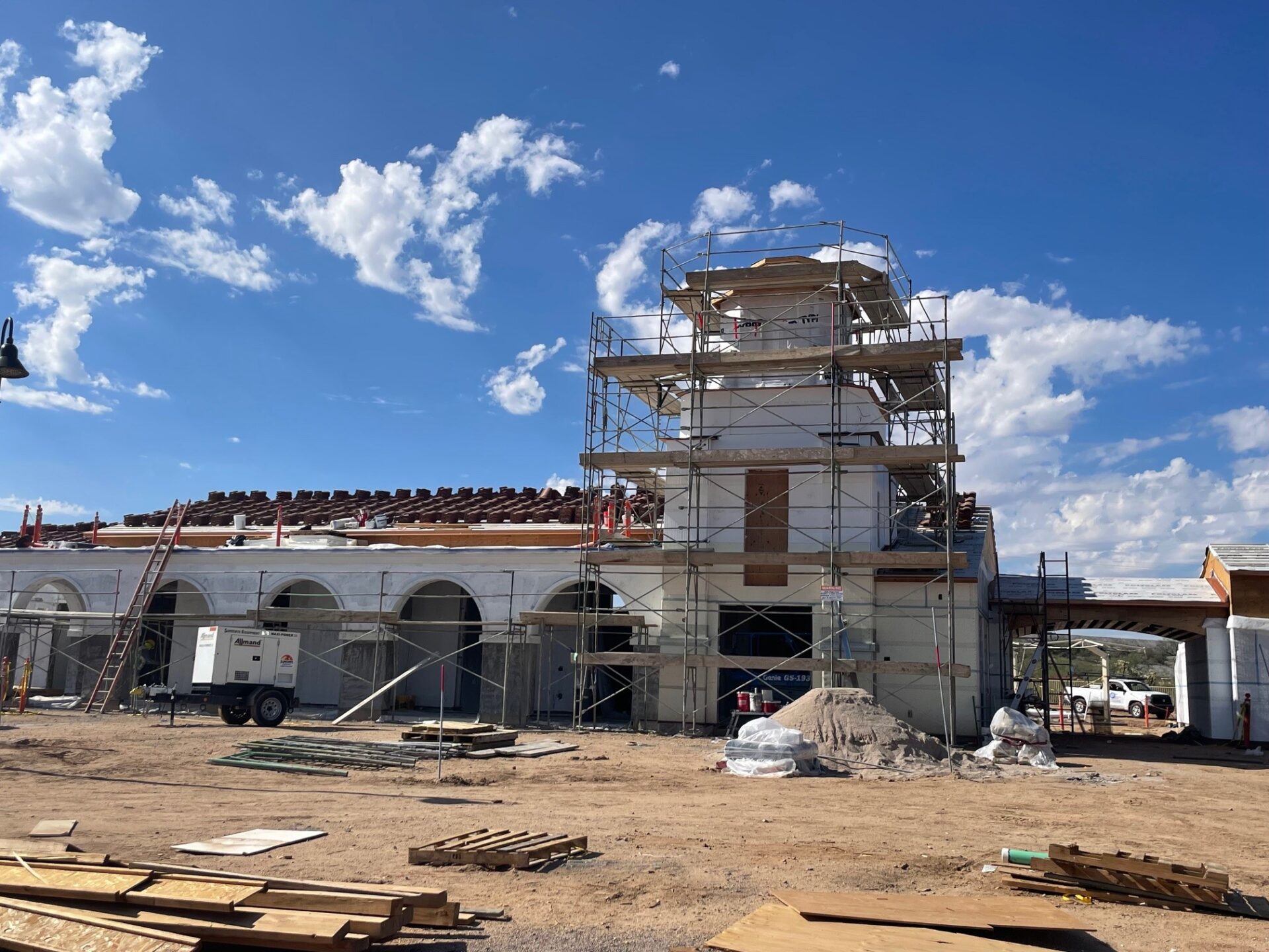 A building under construction with scaffolding surrounding a central tower. Construction materials and equipment are scattered on the dirt ground, and the sky is blue with scattered clouds.