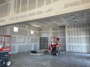 A large indoor space under construction with unfinished drywall on the walls and ceiling, two red scissor lifts, construction materials on the floor, and exposed electrical outlets.