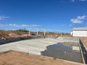 A large, unfinished swimming pool with some water at one end is surrounded by dirt and construction materials under a clear blue sky. A small white building is visible to the right.