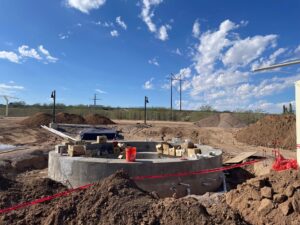 A construction site with a circular concrete structure in the center, surrounded by dirt mounds, electrical wires, and a bright orange bucket. The sky is blue with scattered clouds.